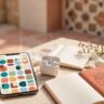 A smartphone, earbuds, blank notebook, and coffee cup sit on a sunny café table.