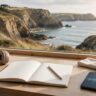 A notebook, headphones, pencil, and phone sit on a desk overlooking the Cornwall coast.