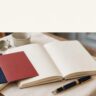 A blank notebook, two colored cards, and a fountain pen on a café table.